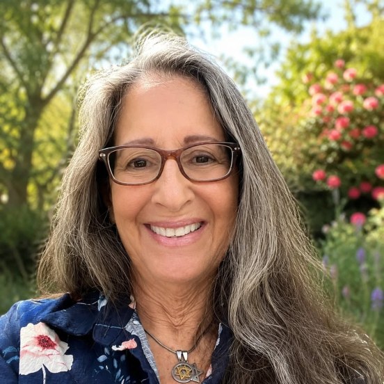 A woman wearing glasses and jeans is smiling at the camera while indoors.